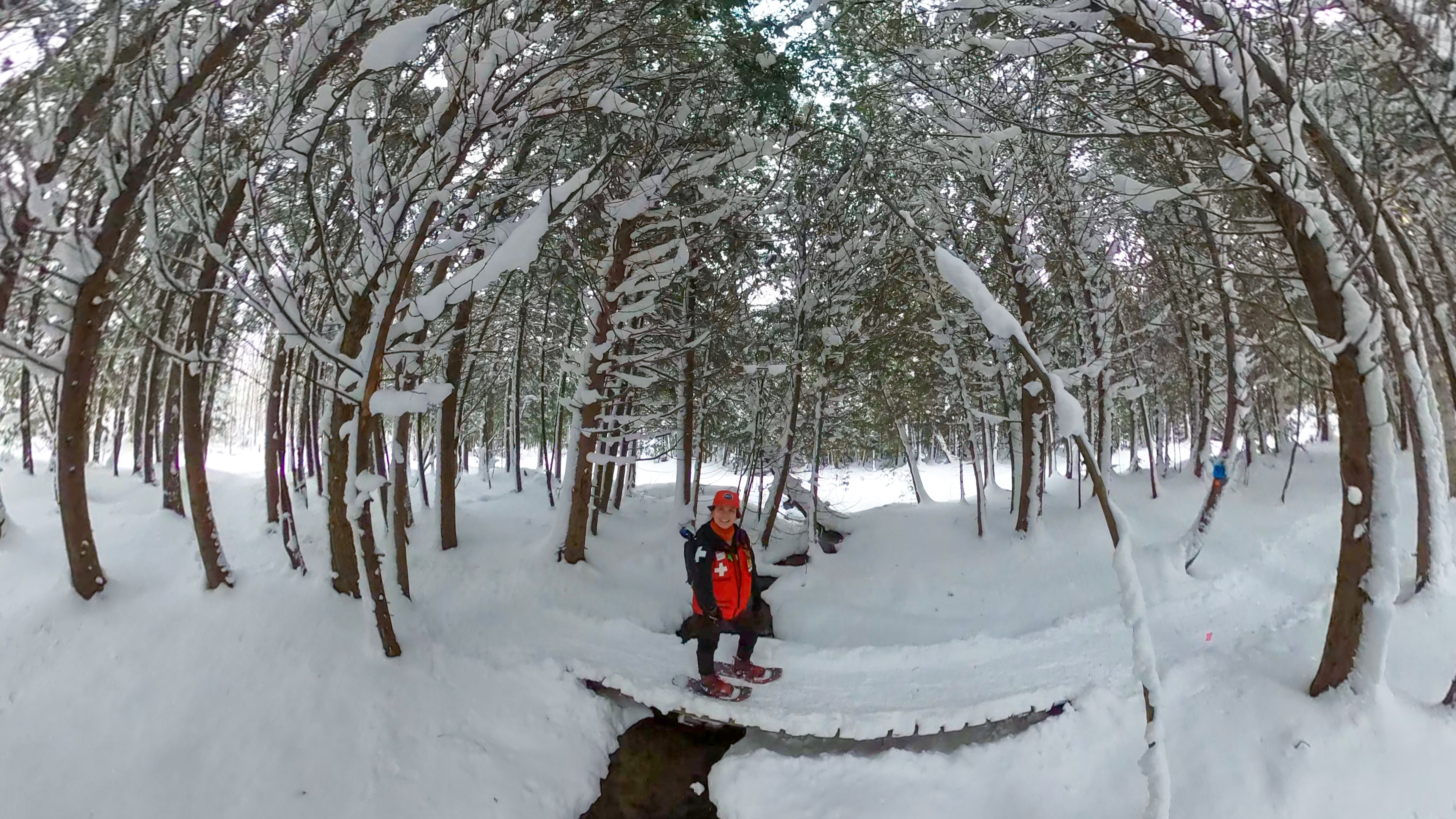 a smiling ski patroller wearing snowshoes stands on a snowy foot bridge in the middle of a forest covered in a fresh blanket of snow.
