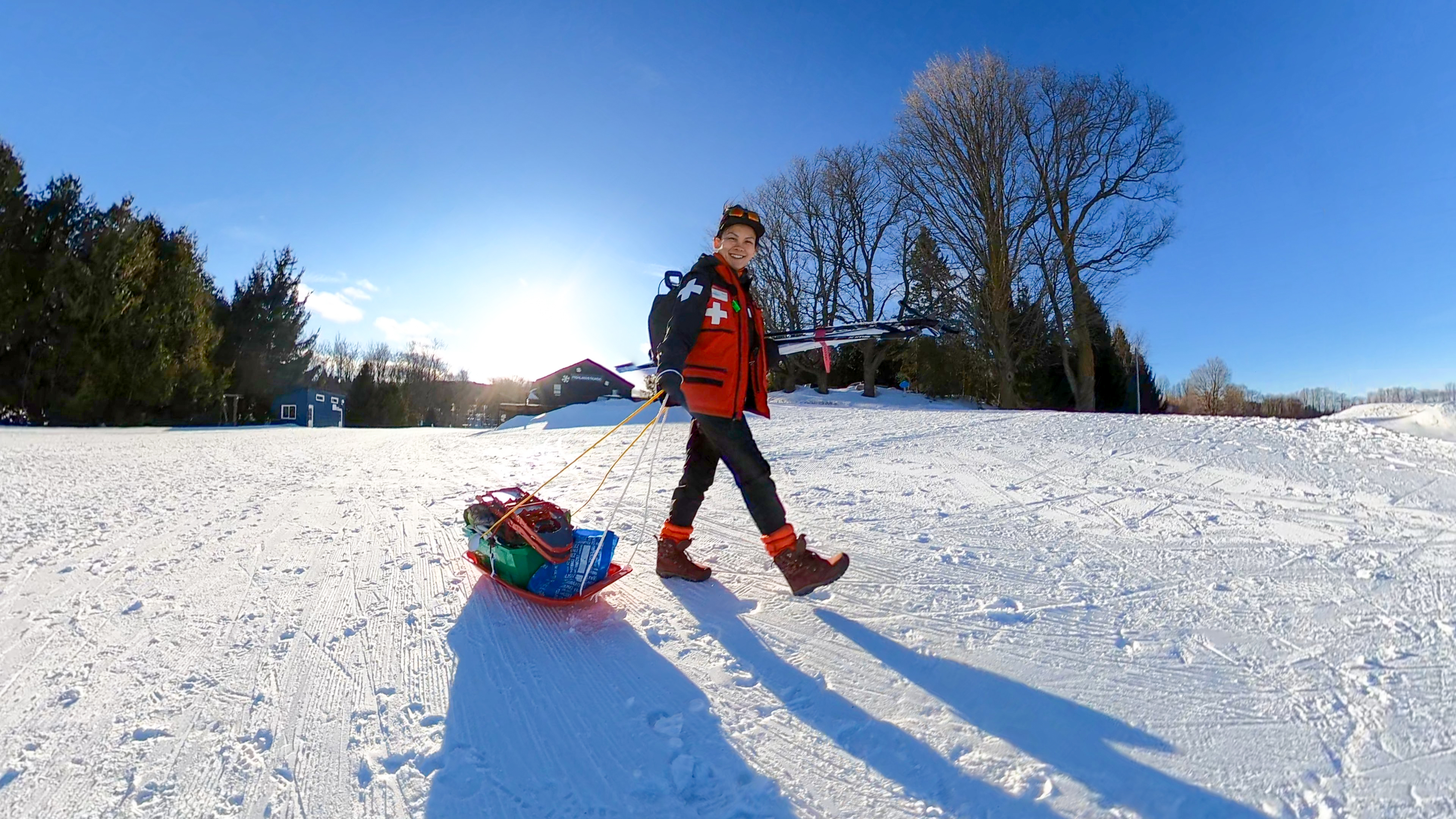 a smiling woman in ski patrol gear pulling a sled loaded with snowshoes and gear across a snowy clearing on a sunny winter day.