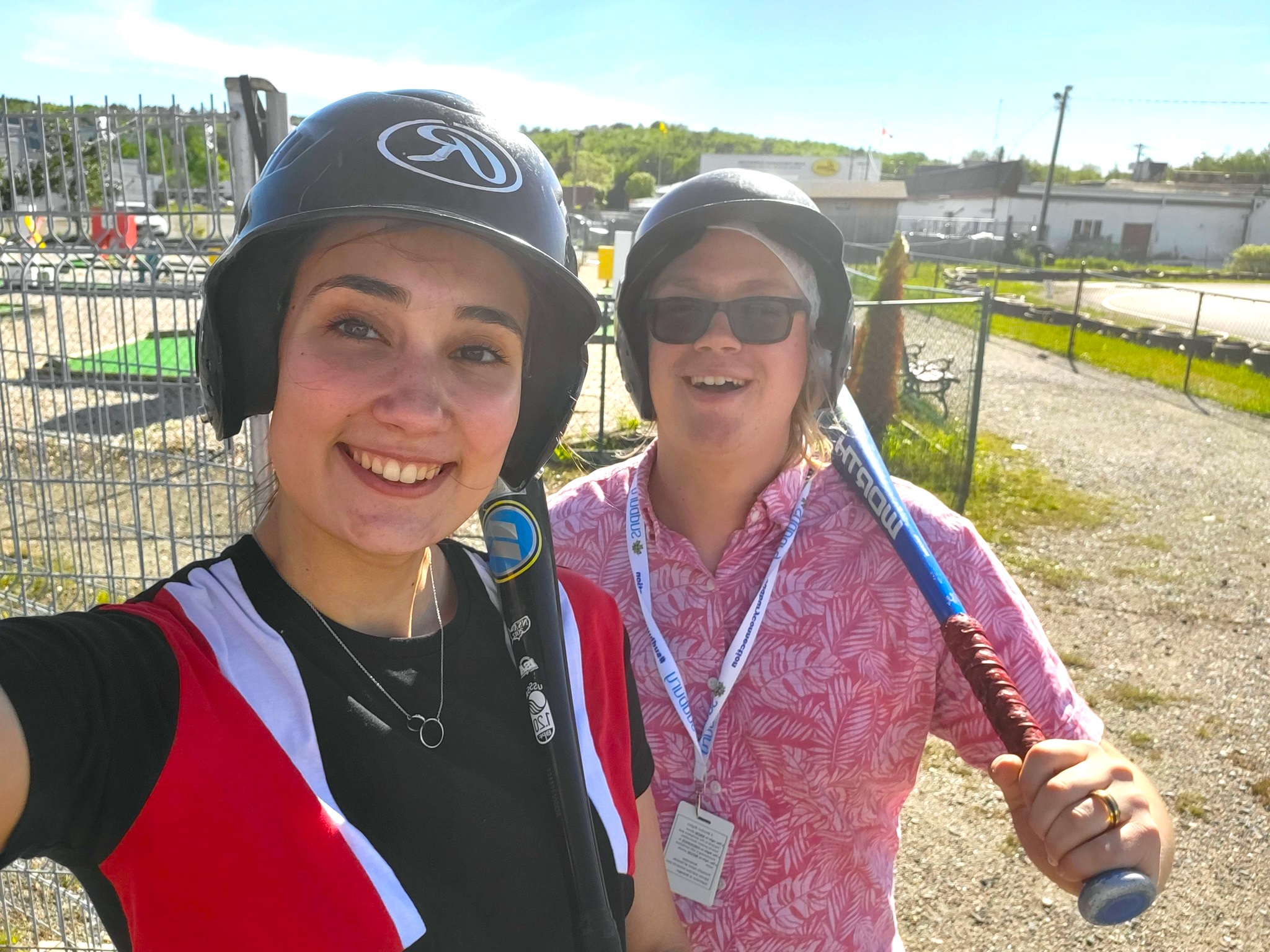 two smiling peple wearing batting helmets and holding bats on a summer day.