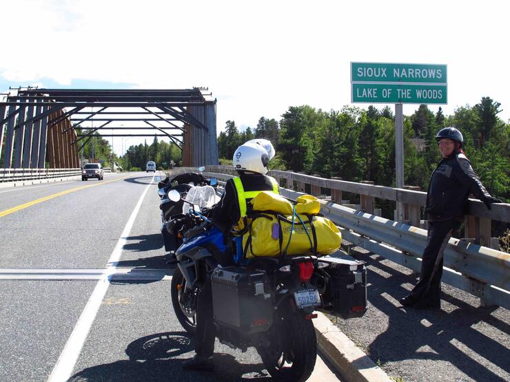 Two motorcyclists parked at the Sioux Narrows/ Lake of the Woods road sign in front of the Sioux Narrows bridge.