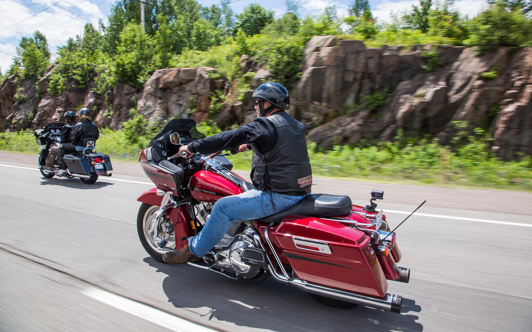 motorcyclists ride down a paved highway next to rocky banks covered in bright green foliage.