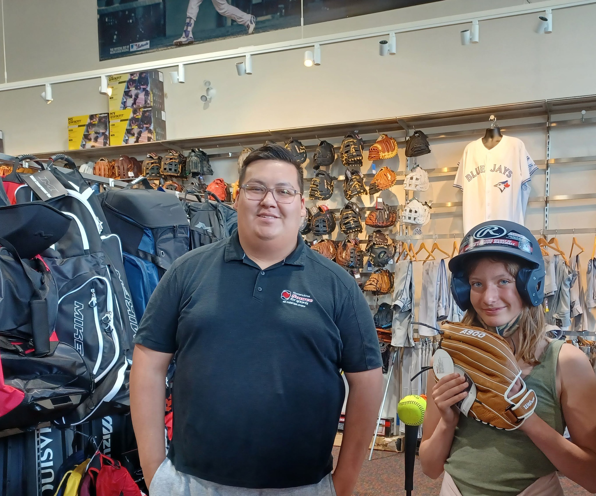 a man in Skater's Edge Source for Sports uniform with a smiling girl who is trying on a baseball helmet and glove.