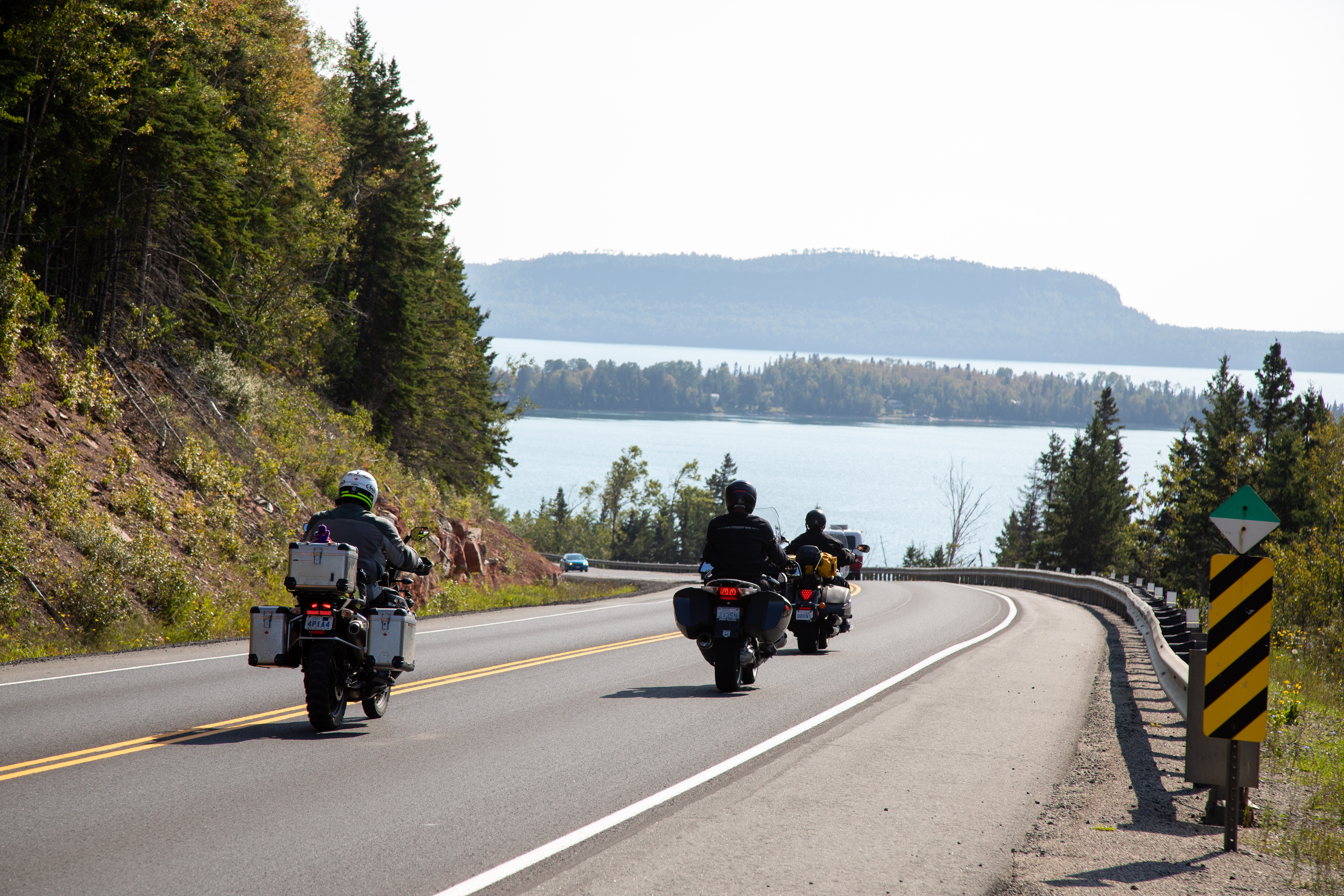 motorcyclists round a bend surrounded by rocky banks and forest, with Lake Superior on the horizon.
