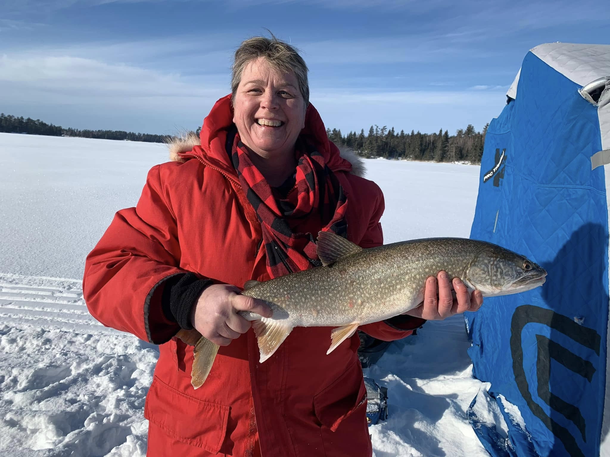 a smiling woman in winter gear holds up a lake trout on a snowy frozen lake.