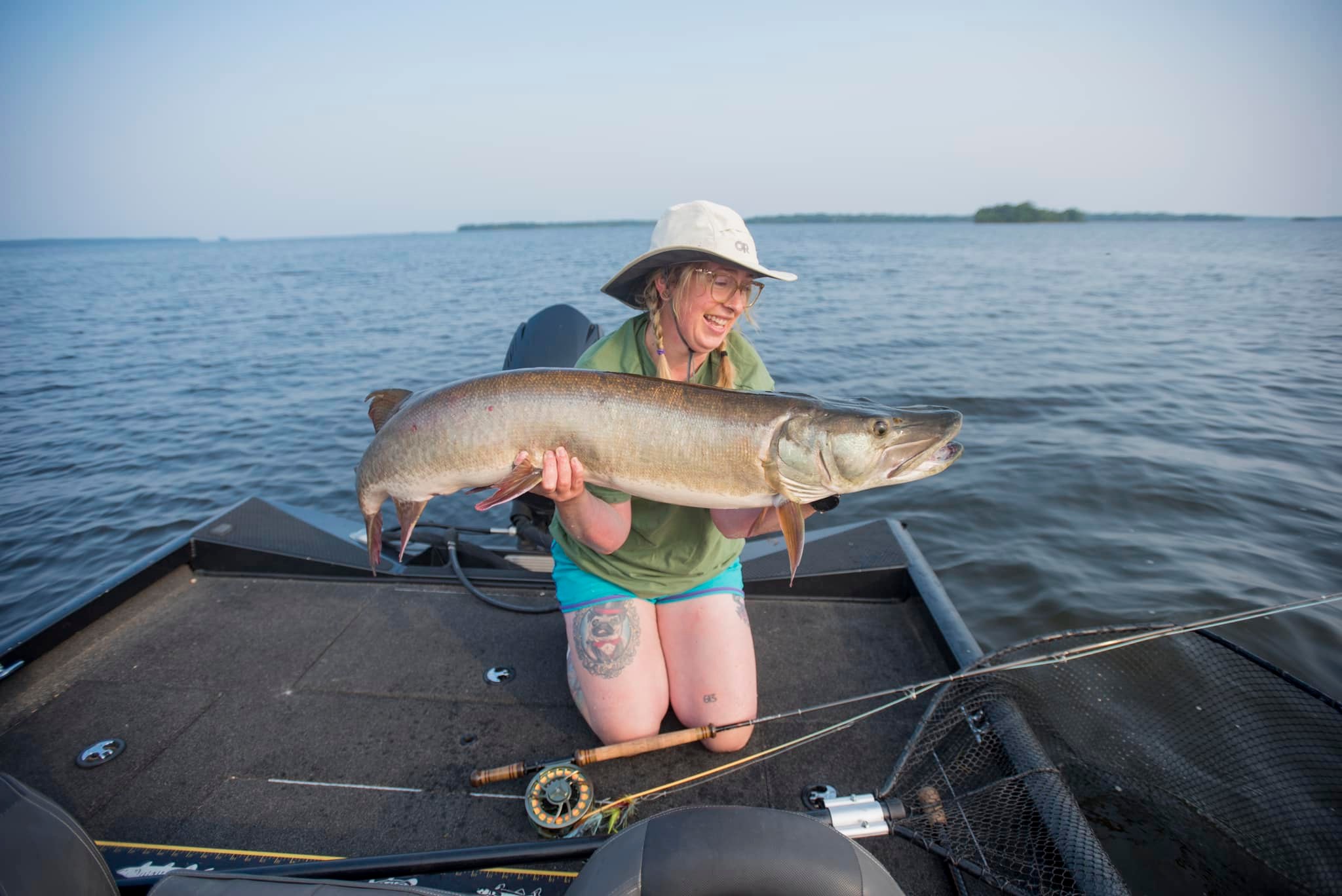 a woman with a fly fishing rod kneels on a boat, smiling and holding up a large muskie.