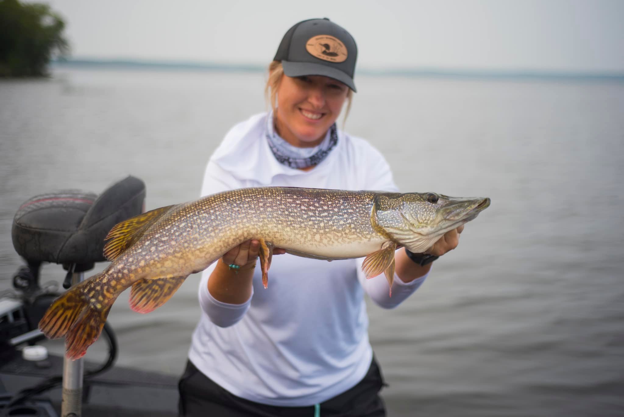 a smiling woman standing on a boat on a smooth lake holds up a large northern pike.