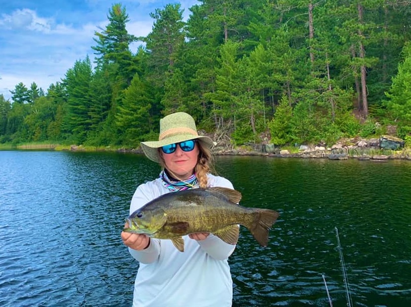 a woman standing next to a lake surrounded in green forest holds up a smallmouth bass.
