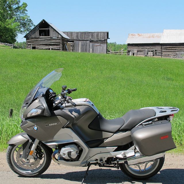 a motorcycle parked next to a green pasture with aged wooden farm buildings on a sunny spring day.