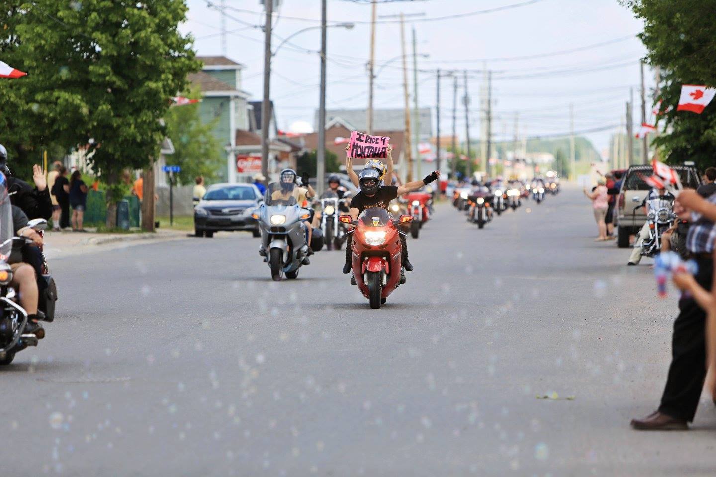 motorcyclists ride through New Liskeard amidst a crowd of cheering people waving Canada flags and blowing bubbles on a summer day. One rider holds up a sign that reads "I ride for Montana".