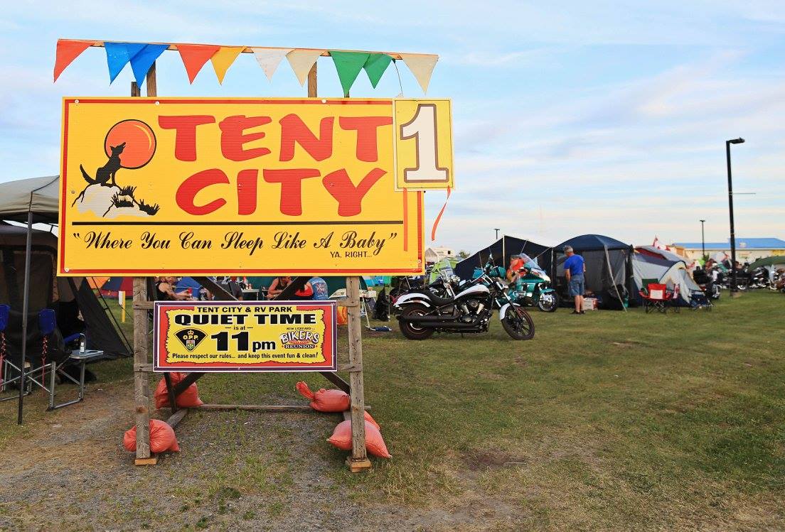 a large wooden sign that reads "Tent City, Where You Can Sleep Like A Baby" on a grassy field with tents and motorcycles in the background.