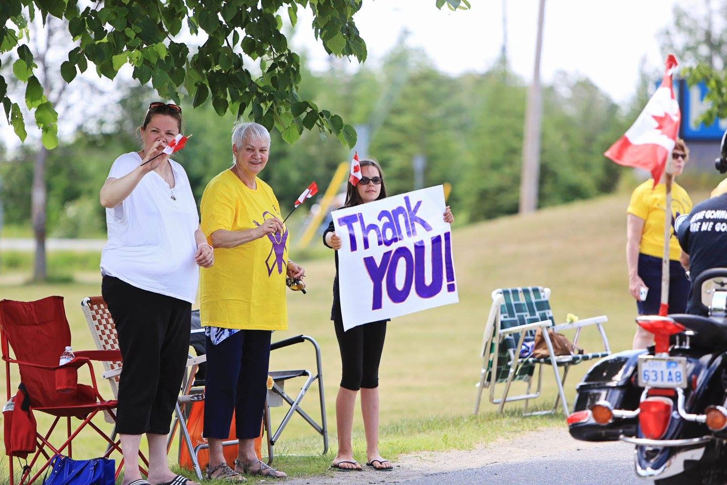 people watching the Bikers Reunion parade on a summer day wave Canada flags and a sign that reads "Thank You!"