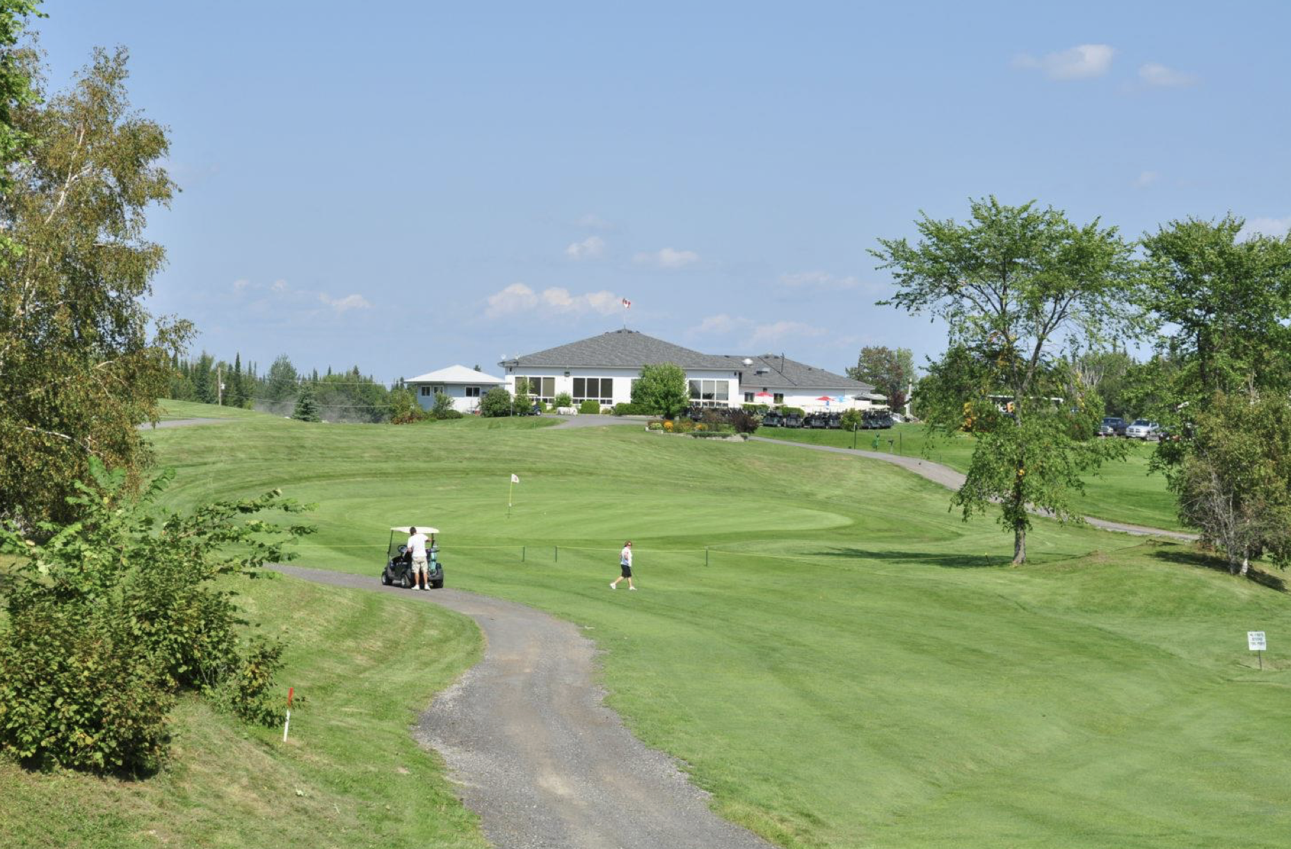golfers play on a summer day, on a rolling green course dotted with trees and a large clubhouse in the distance.