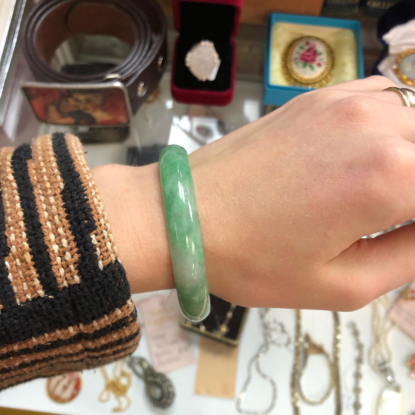 a hand wearing a vintage jade bangle, held up above a table laid with rows of beautiful assorted vintage jewellery.
