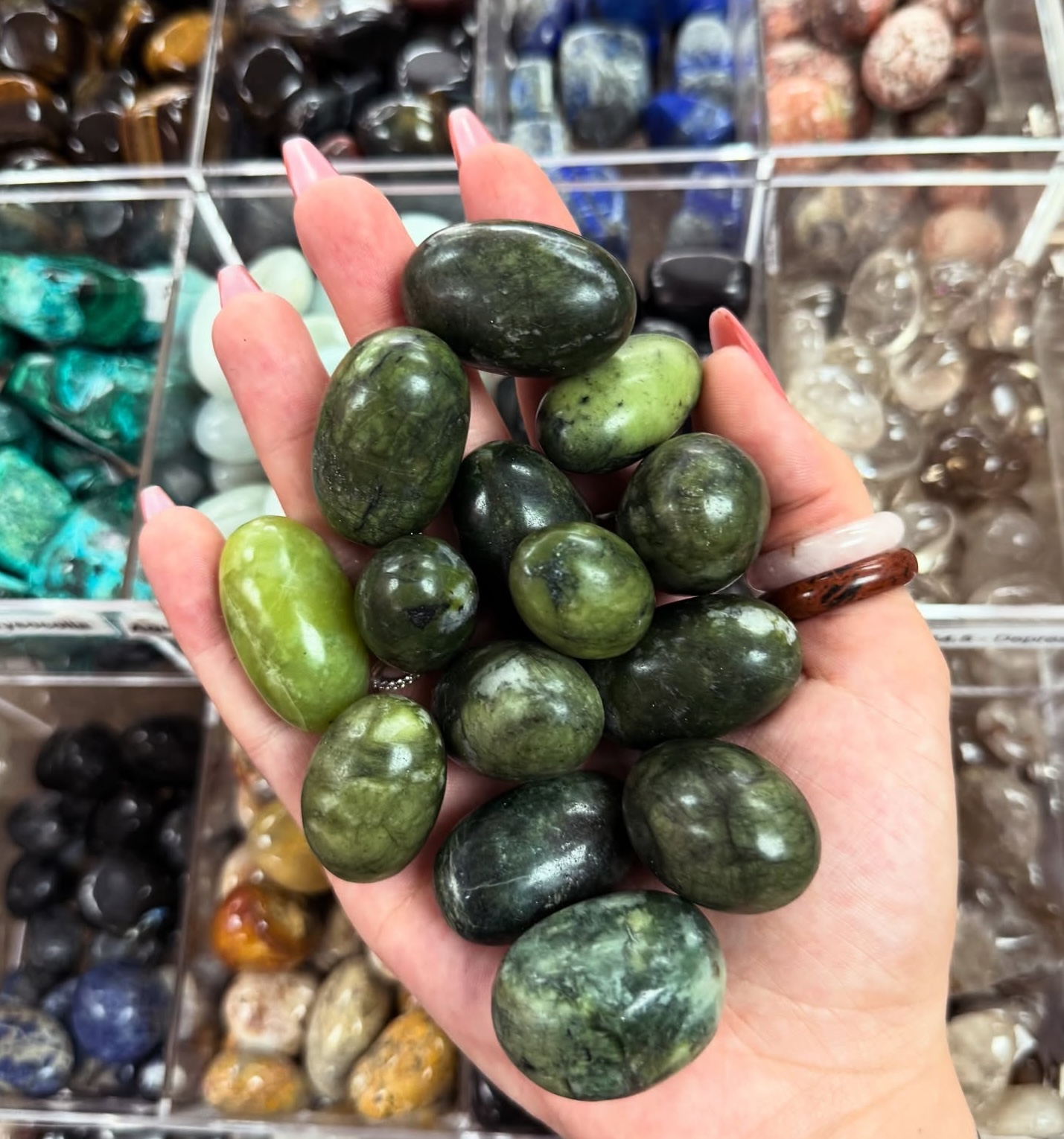 a hand filled with polished green gemstones, with shelves of multicoloured stones in the background.