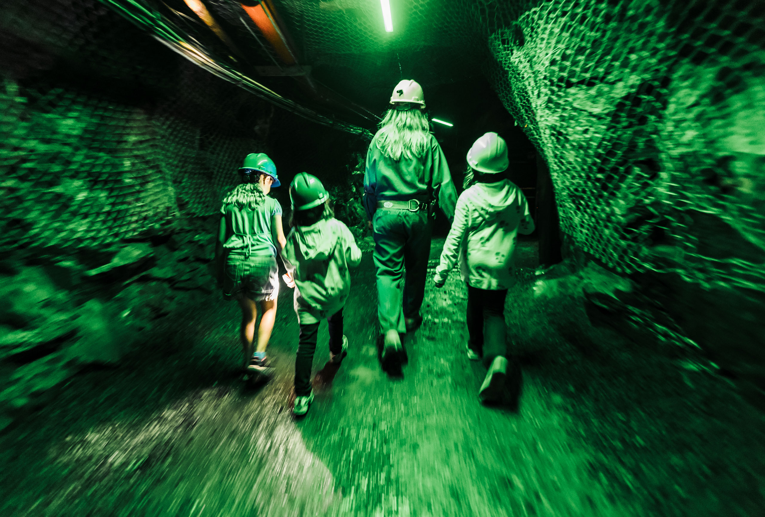 a group of children walk with a guide through a rock tunnel, illuminated by green lighting. All are wearing mining helmets.