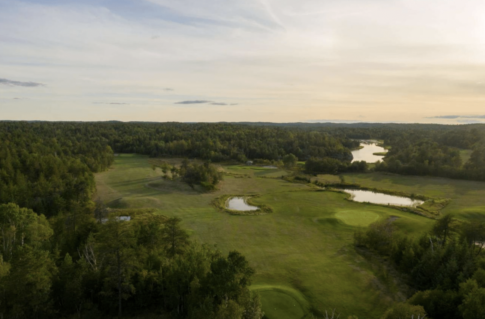 a smooth green golf course surrounded by lush forest and dotted with water hazards under a soft golden sunset.