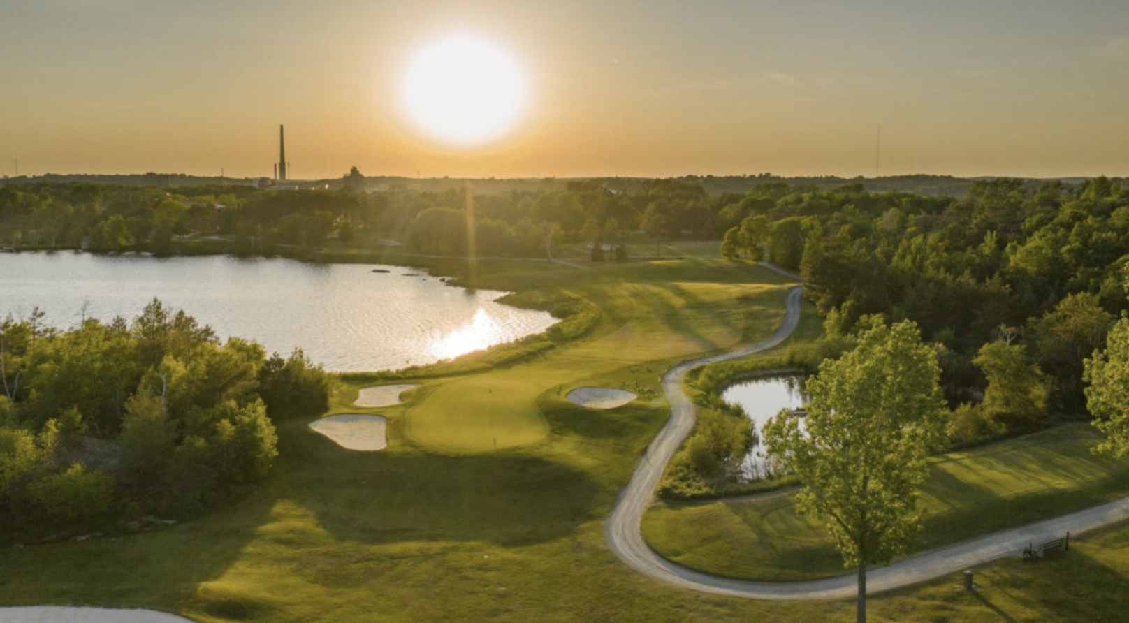 A large rolling golf green next to a shining lake bathed in a golden sunset.