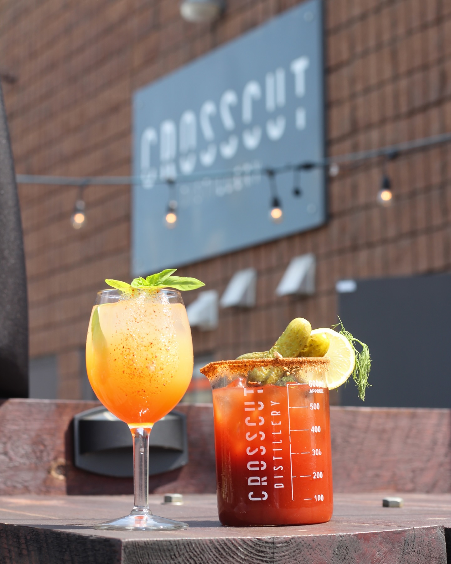 two cocktails garnished with lemon and herb sprigs. A Crosscut Distillery sign is in the background.