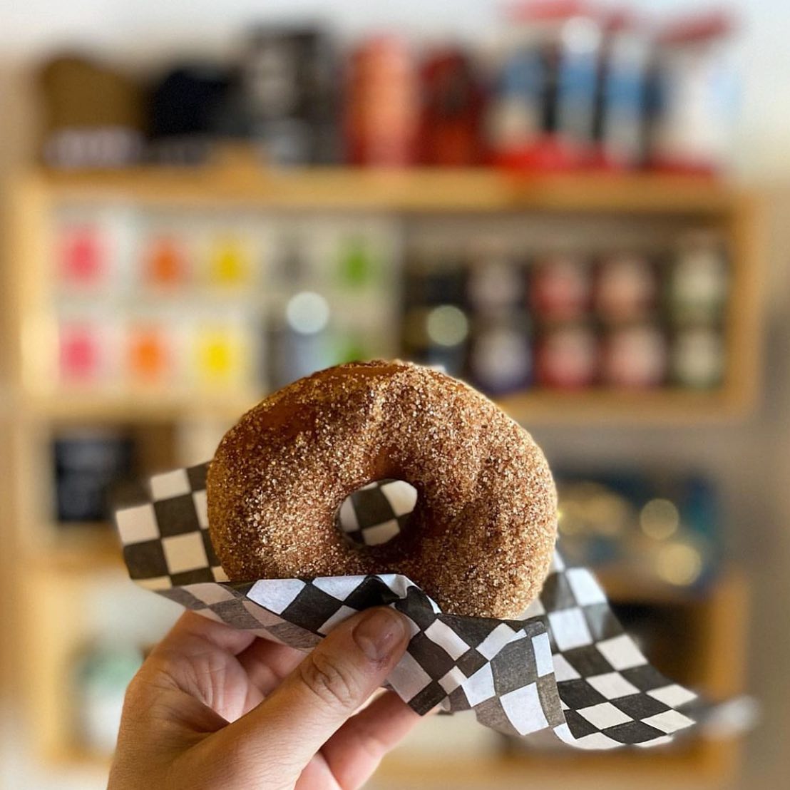 a hand holds up a donut wrapped in checkered paper in front of shelves of spices and jams.
