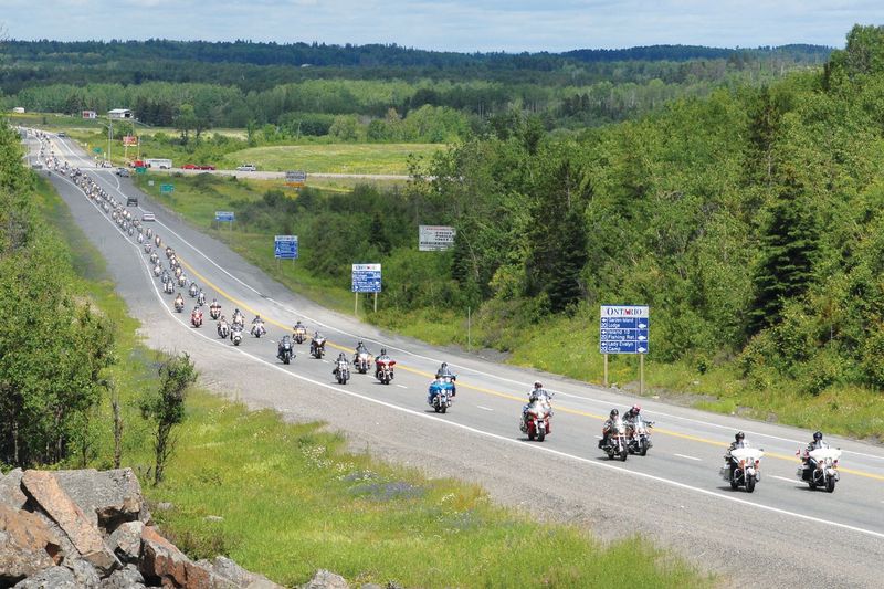 a long parade of motorcycles stretching down the highway to the horizon on a summer day, surrounded by green trees and rolling grassy fields.
