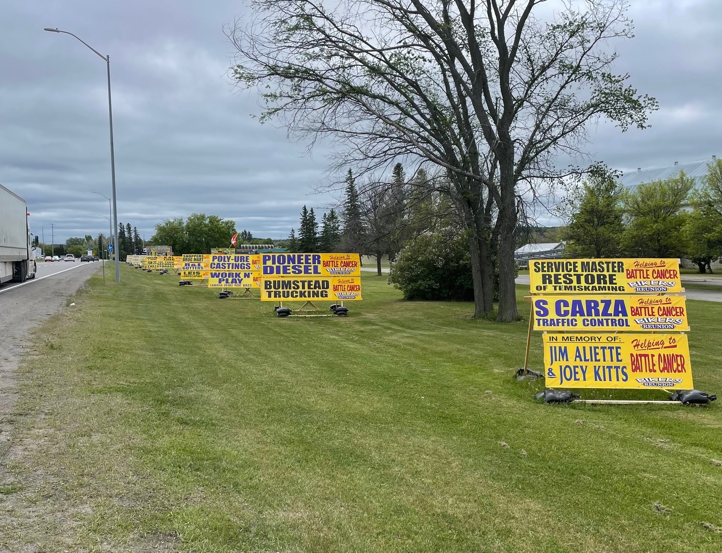 a row of large yellow signs in the green grass along the side of a highway, stretching off into the distance. The signs are brightly labelled with the names of businesses and families that have sponsored the New Liskeard Bikers Reunion in memory of a loved one.