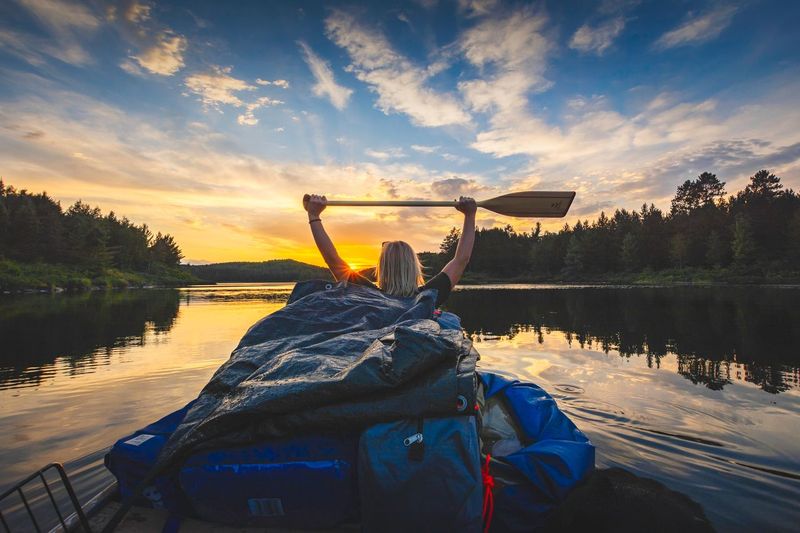 A person sitting in a canoe filled with camping gear raises their paddle above their head as they float on a glassy lake at sunset in the summer.