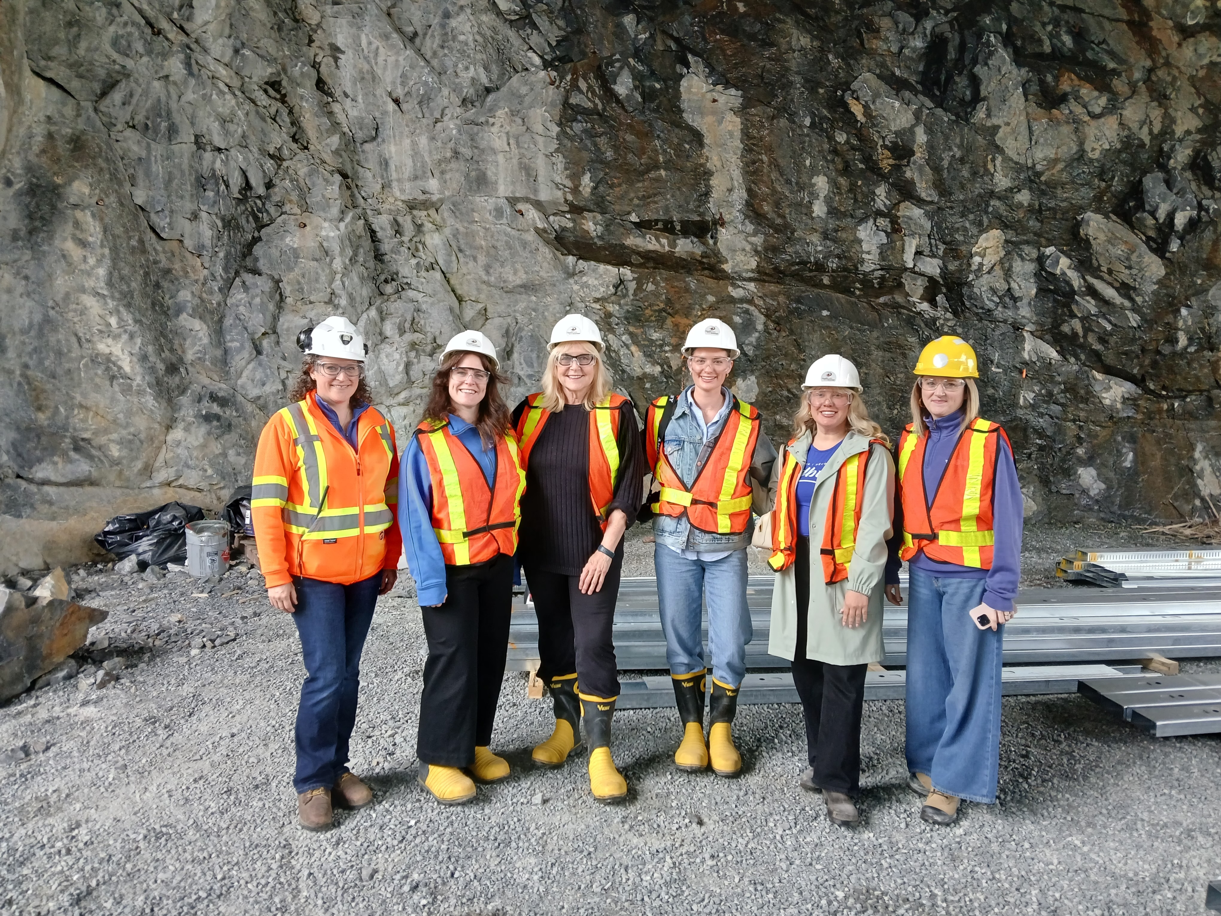 Susan Truppe poses with several other representatives of Sudbury and Dynamic Earth, all wearing hard hats and reflective vests amd smiling warmly. They are standing in a rock cave.