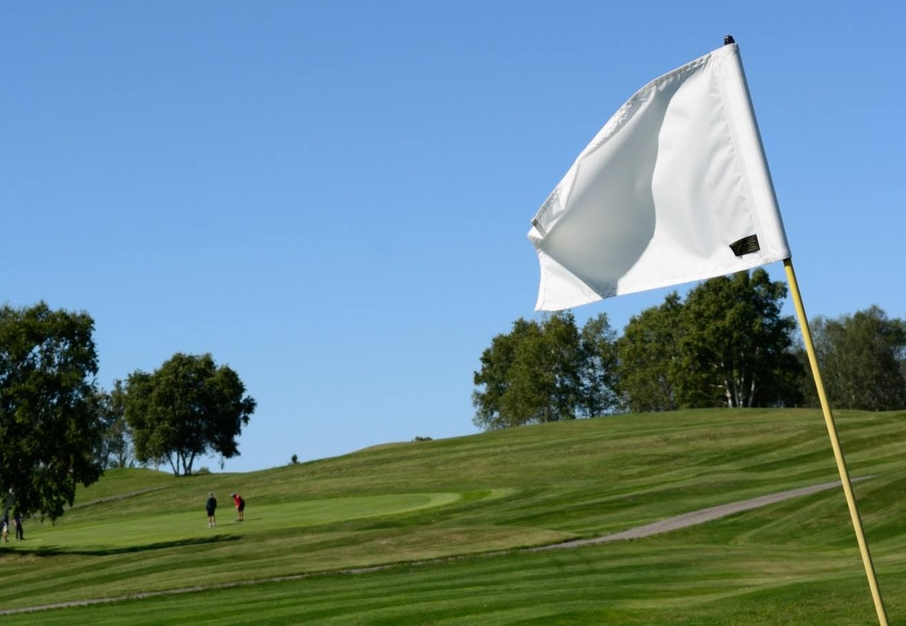 a golf flag flying above the green on a clear summer day, golfers playing in the background.