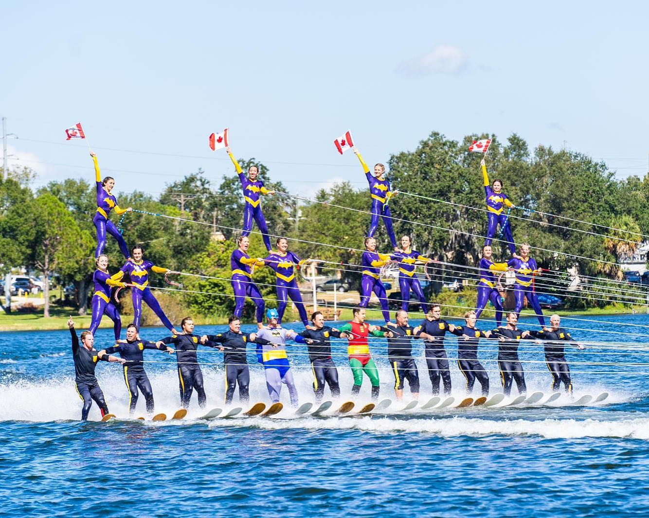 waterskiers in superhero costumes glide across a lake in a 3-tier pyramid on a bright summer day.