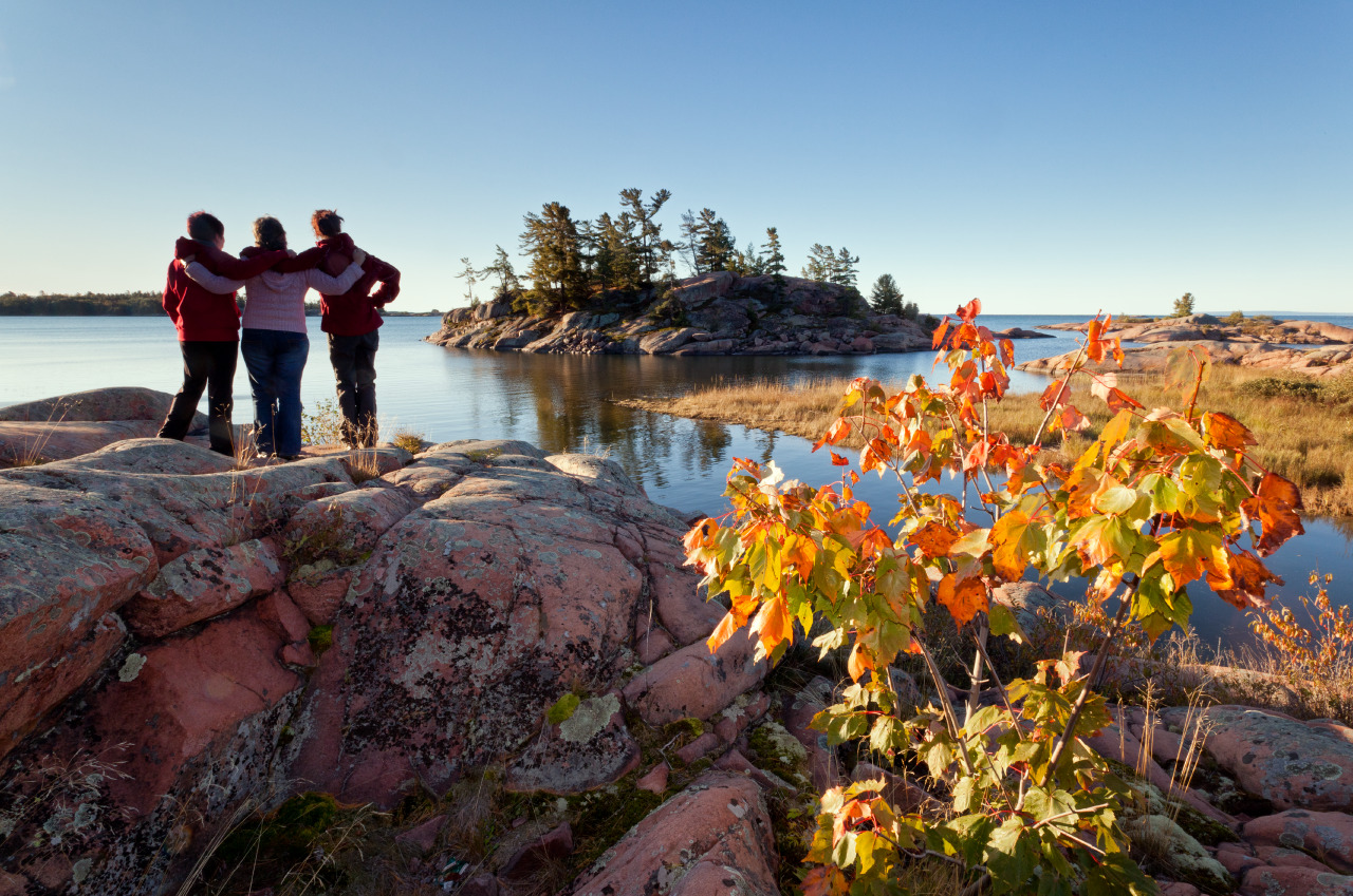 3 people stand with their arms over each others shoulders, looking out over a shining lake surrounded by fall foliage.