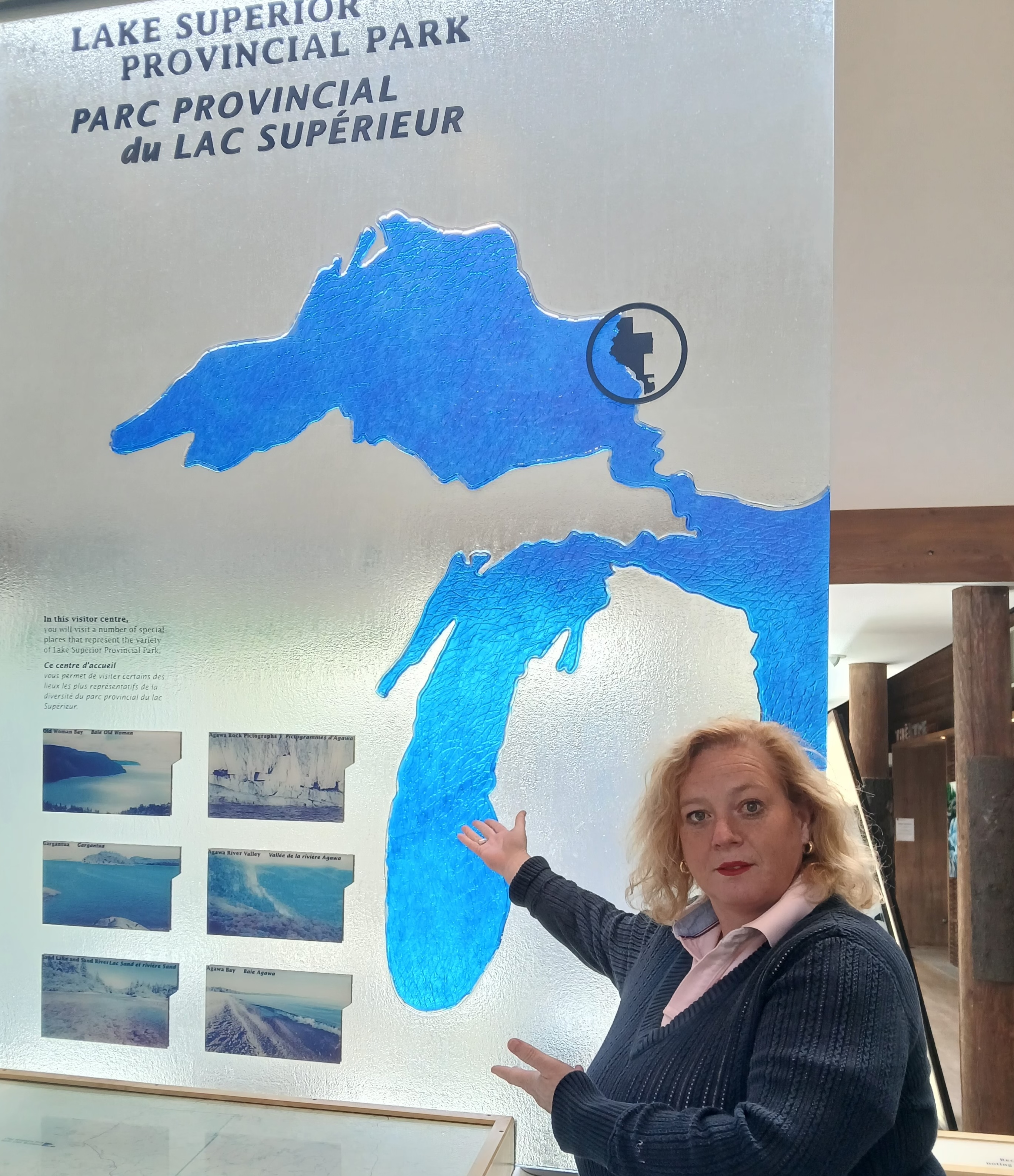 Lisa MacLeod poses with an expression of mock-seriousness, her hands up and gesturing to a large informational plaque behind her that reads "Lake Superior Provincial Park" and shows the Great Lakes.