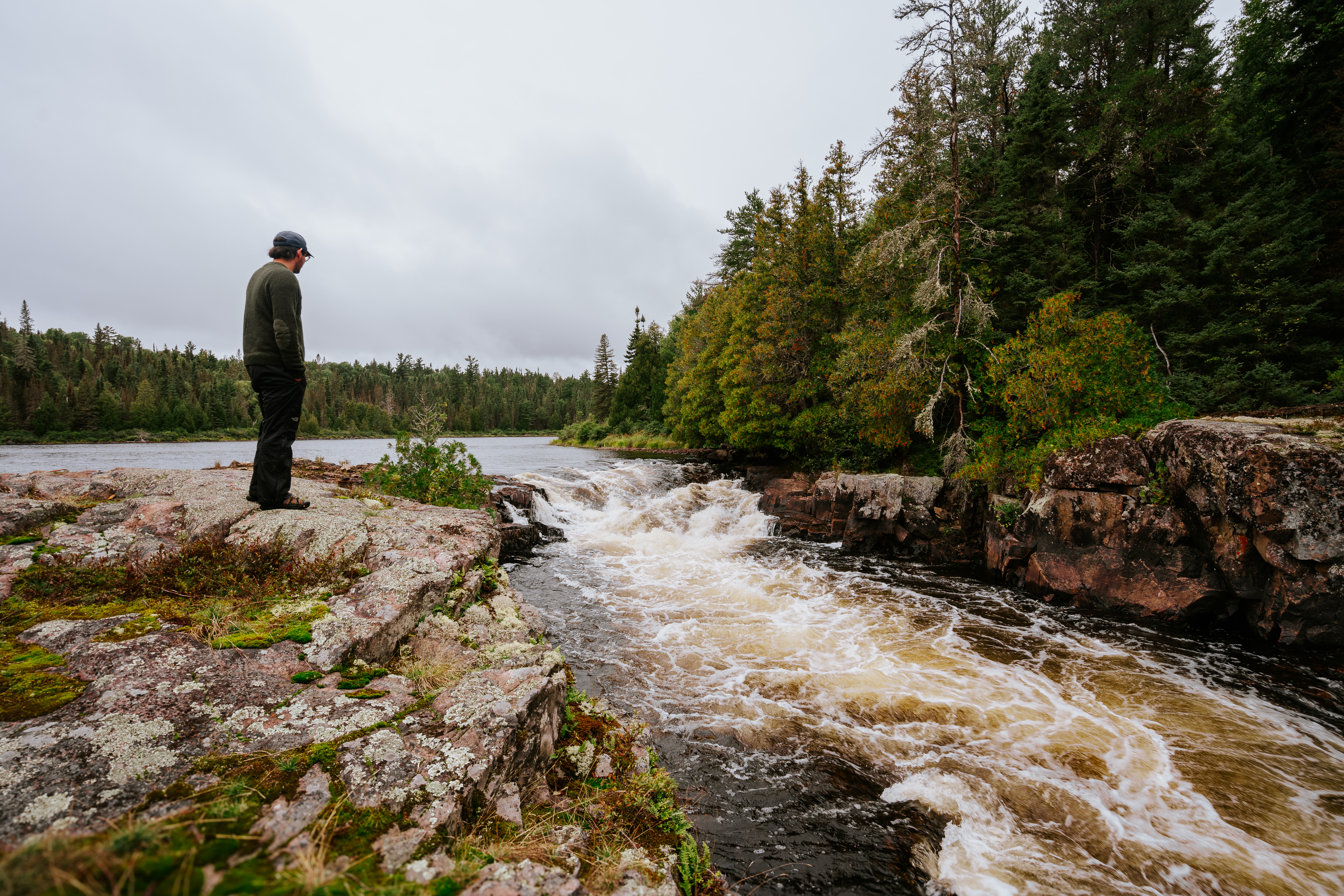 a person looks at the rocky rapids on the Spanish River, surrounded by green wilderness. 