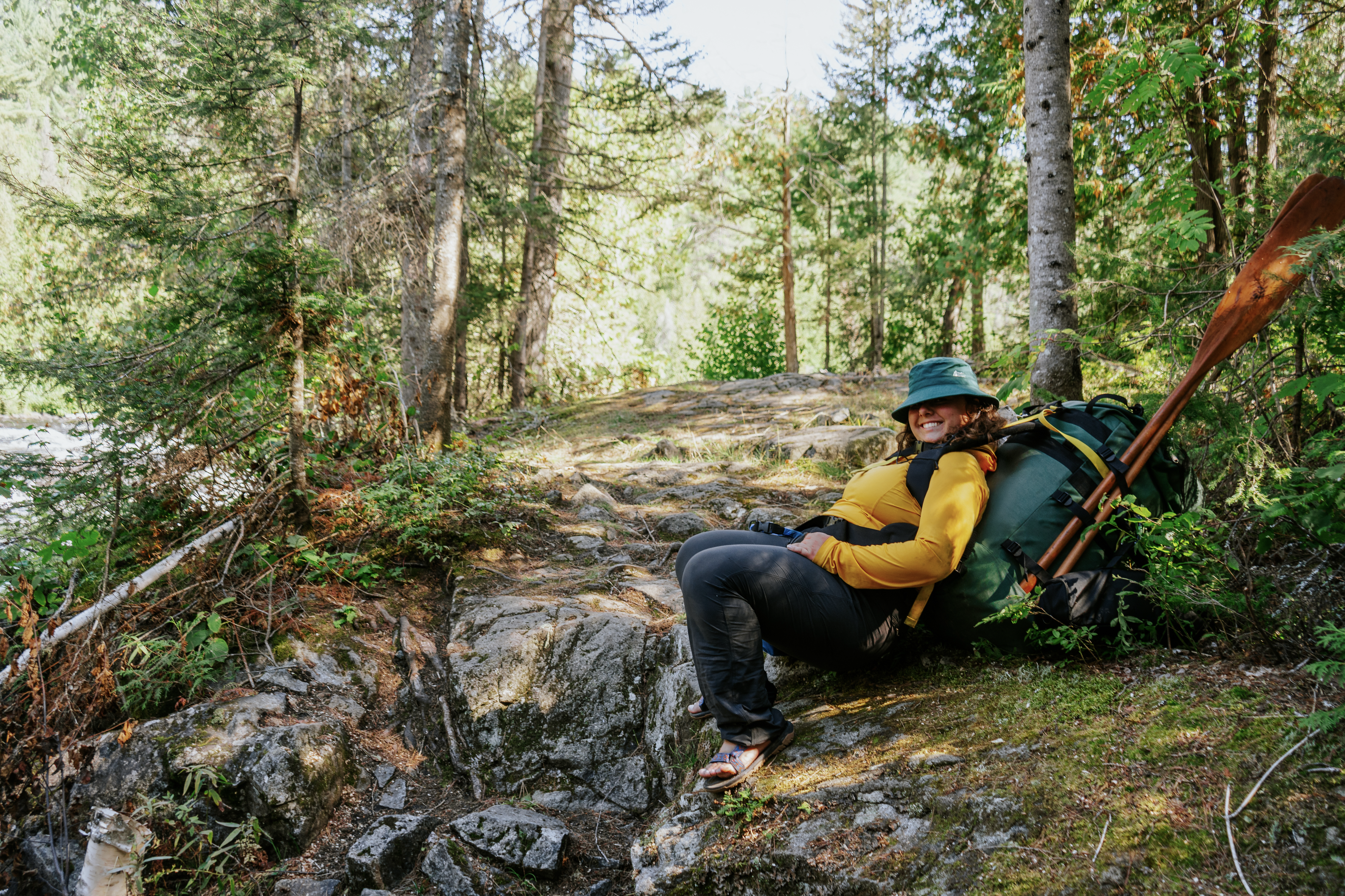 a smiling woman wearing a hat and a large hiking pack rests with her canoe paddle on a mossy, forested river bank on the Spanish River.