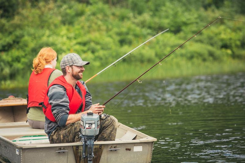 a man and woman fish happily from a boat on a lake surrounded by green forest.