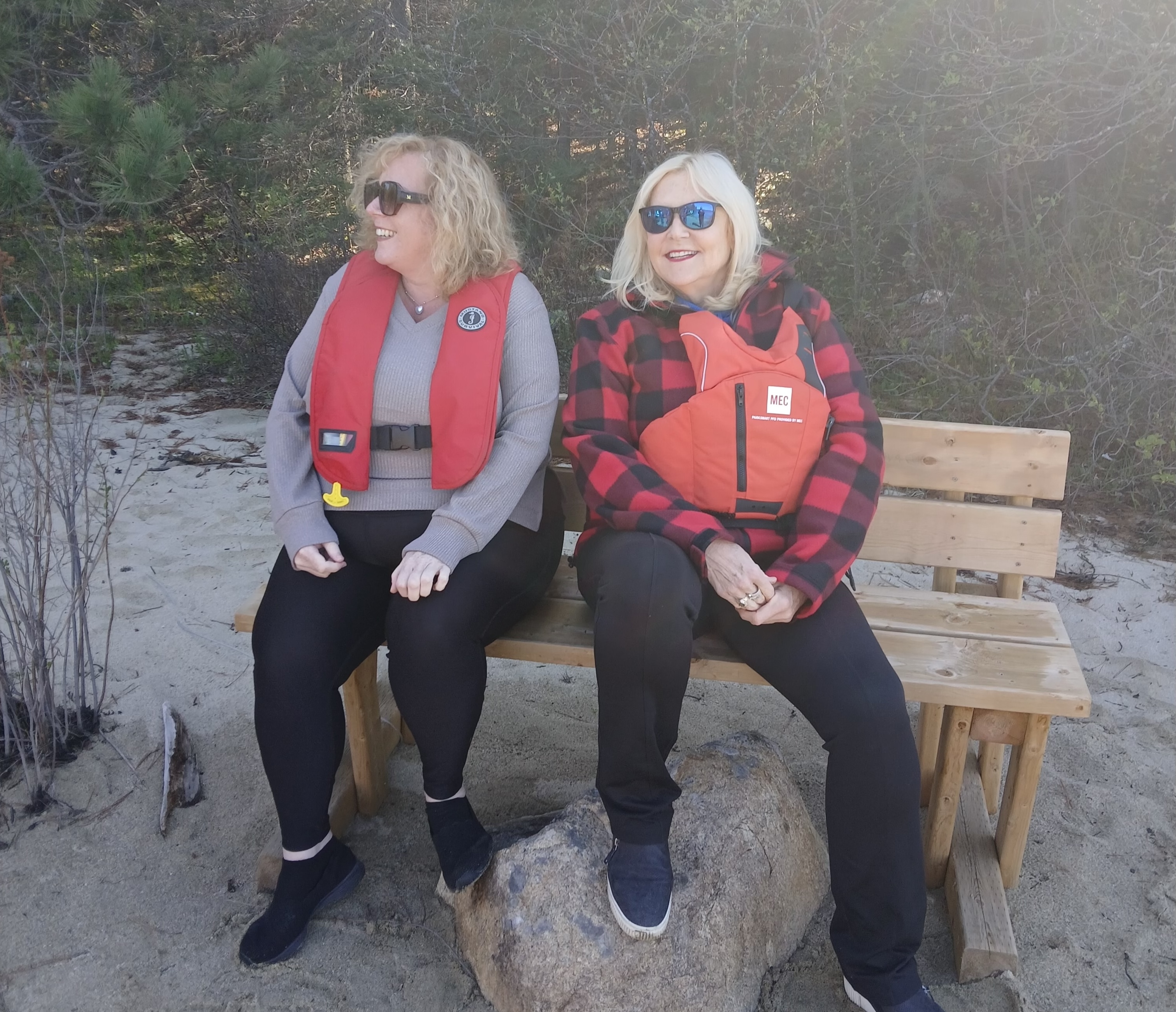 Susan Truppe and Lisa MacLeod smile on a bench on a sunny beach next to green boreal forest. They are wearing life jackets and sunglasses.