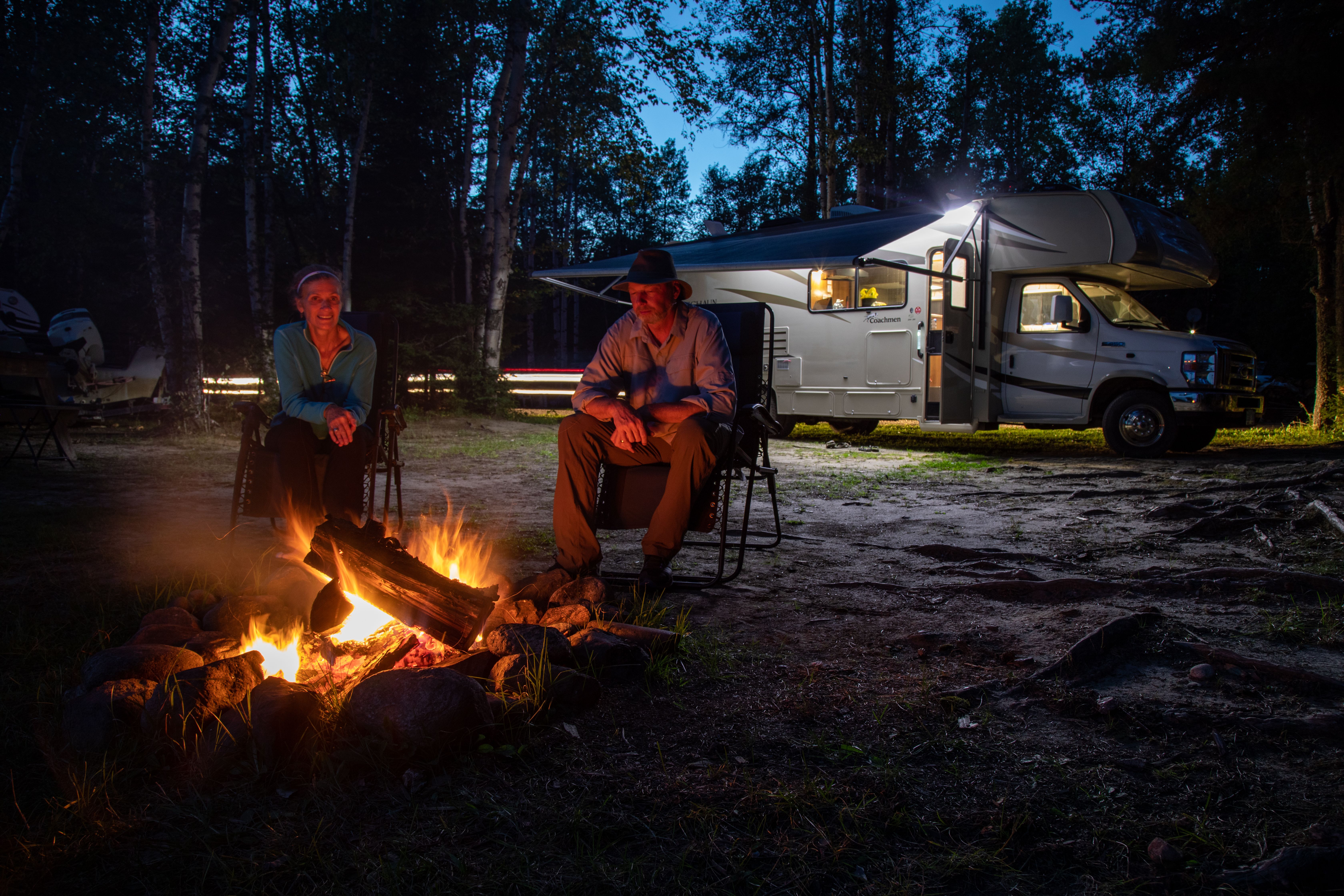 a man and woman sit by a campfire in the evening, next to their RV in the forest.