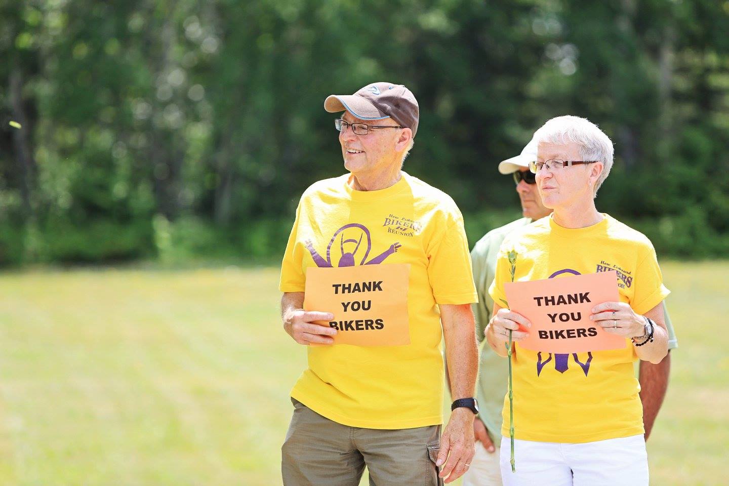 an elderly man and woman wearing yellow Bikers Reunion t-shirts smile as they stand outside on a summer day next to green forest. They are holding paper signs that say "Thank You Bikers" and looking into the distance.