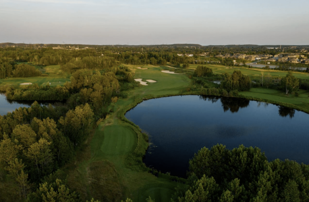 A wide green, forested golf course circling around a glassy lake.