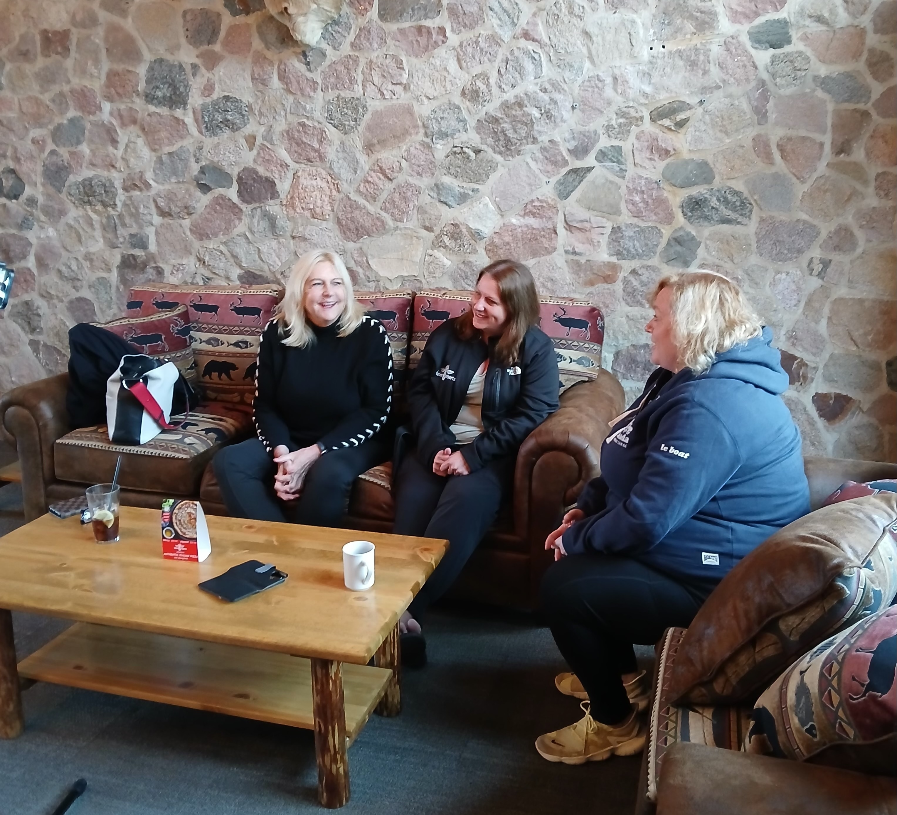 Lisa MacLeod and Susan Truppe talk with a representative of Totem Resorts in Kenora, smiling as they chat together on couches in front of the lodge's rustic stone and mortar walls.