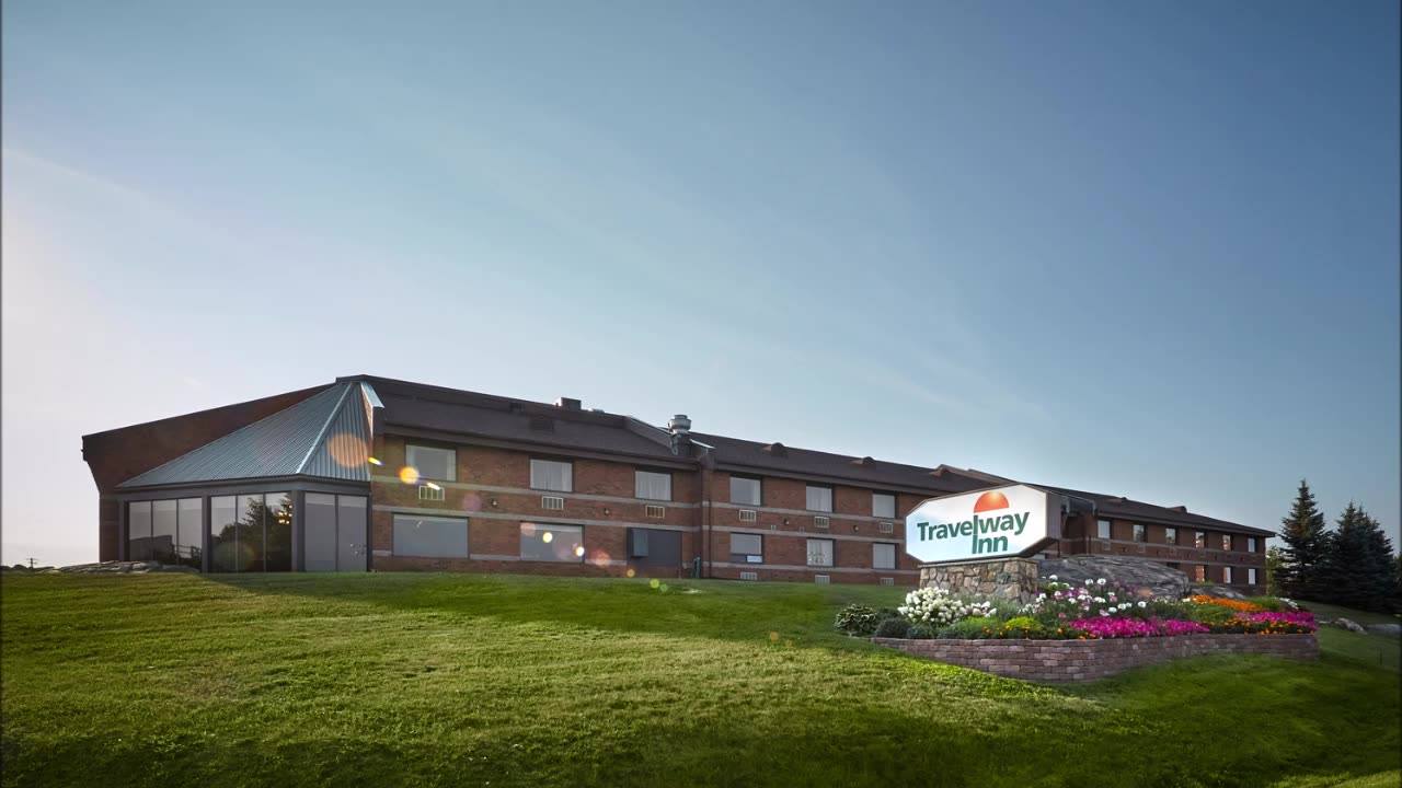 the Travelway Inn Sudbury on a lush green rolling lawn under a clear blue sky.