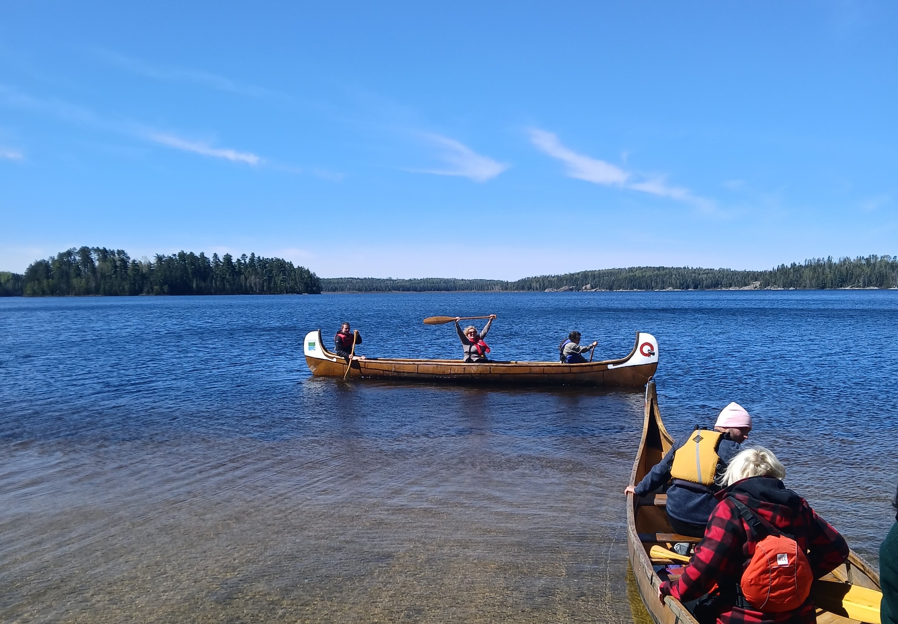 A woman in a canoe with two other people holds her paddle high above her head triumpantly as she smiles, part of a party of two canoes full of paddlers on a clear shining lake on a summer day.