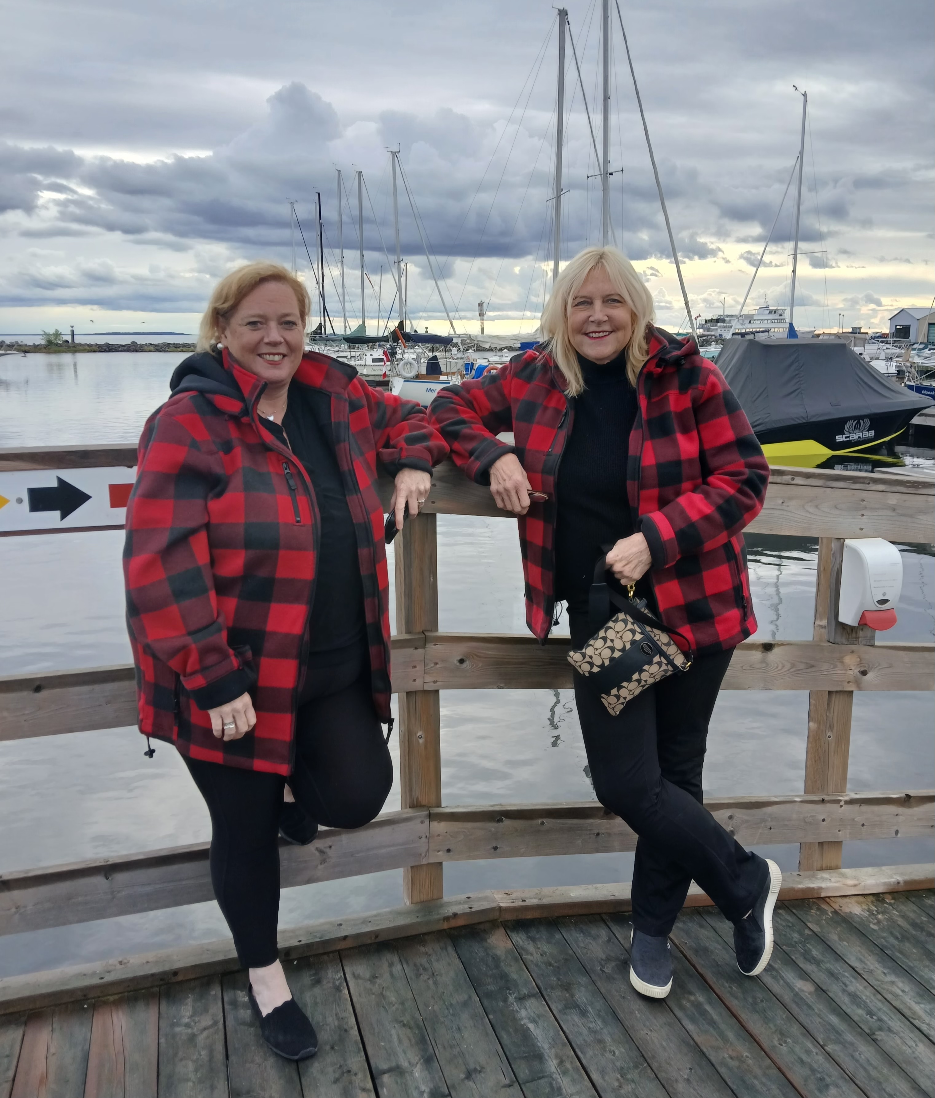 Susan Truppe and Lisa MacLeod smile as they stand in front of a harbour in Moosenee Ontario on a cloudy, breezy day.