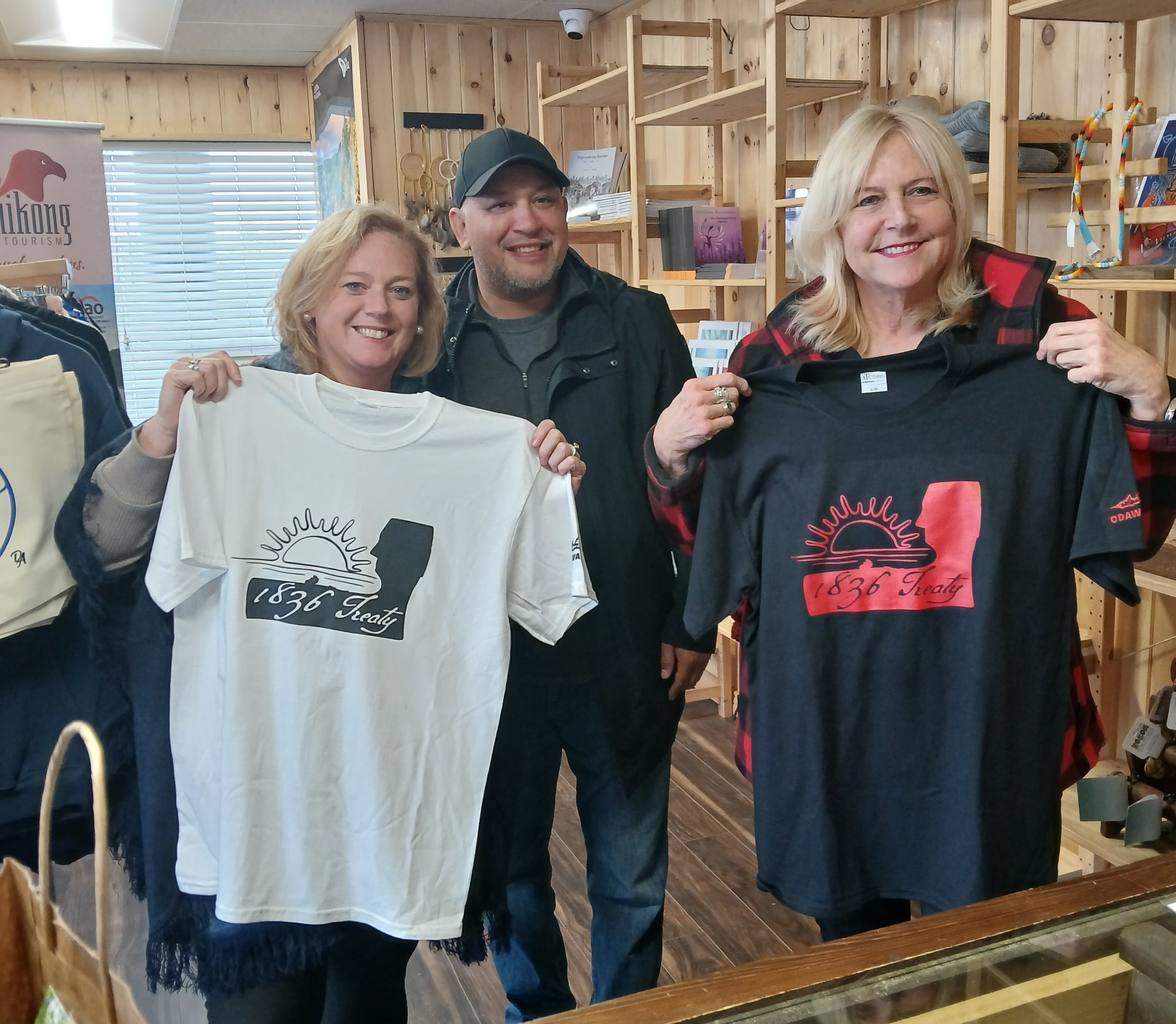 Lisa MacLeod and Susan Truppe smile as they hold up t-shirts printed with a silhouette of a person's face, a sun and the words "1836 Treaty". They are standing in a shop in Wikwemikong First Nation. Their host smiles behind them.