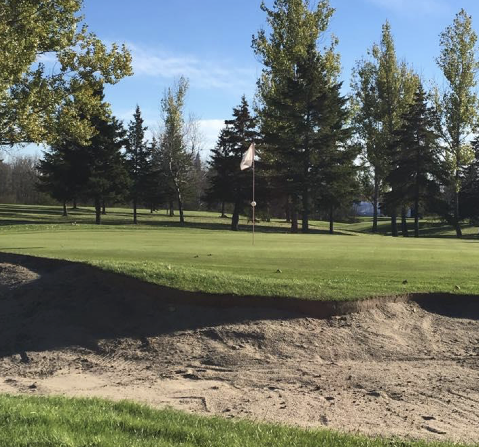 a lush golf course with a soft sand trap and green trees throughout on a summer day.