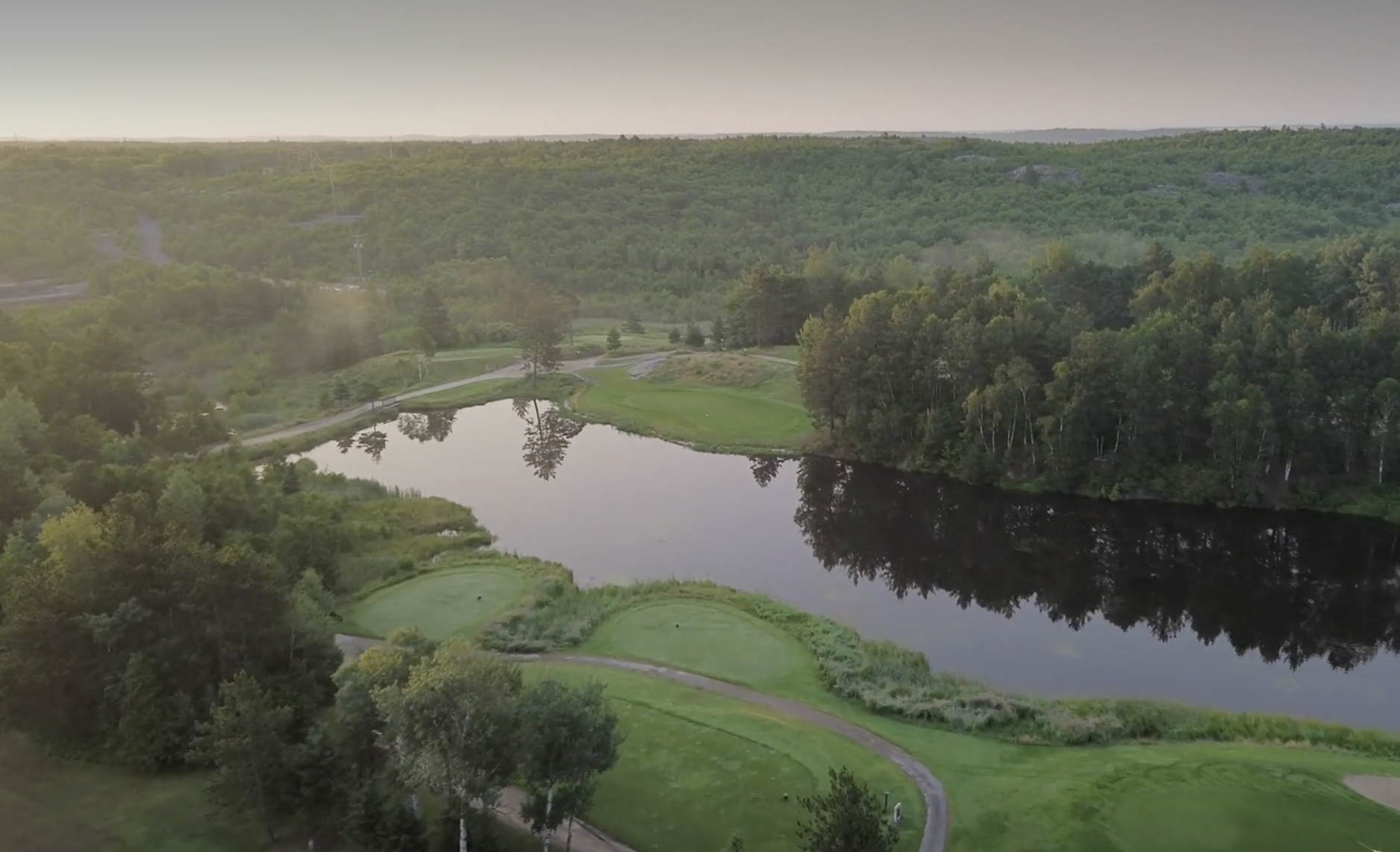 a lush green golf course on a misty summer morning, surrounded by forest, with a shining pond in the centre.