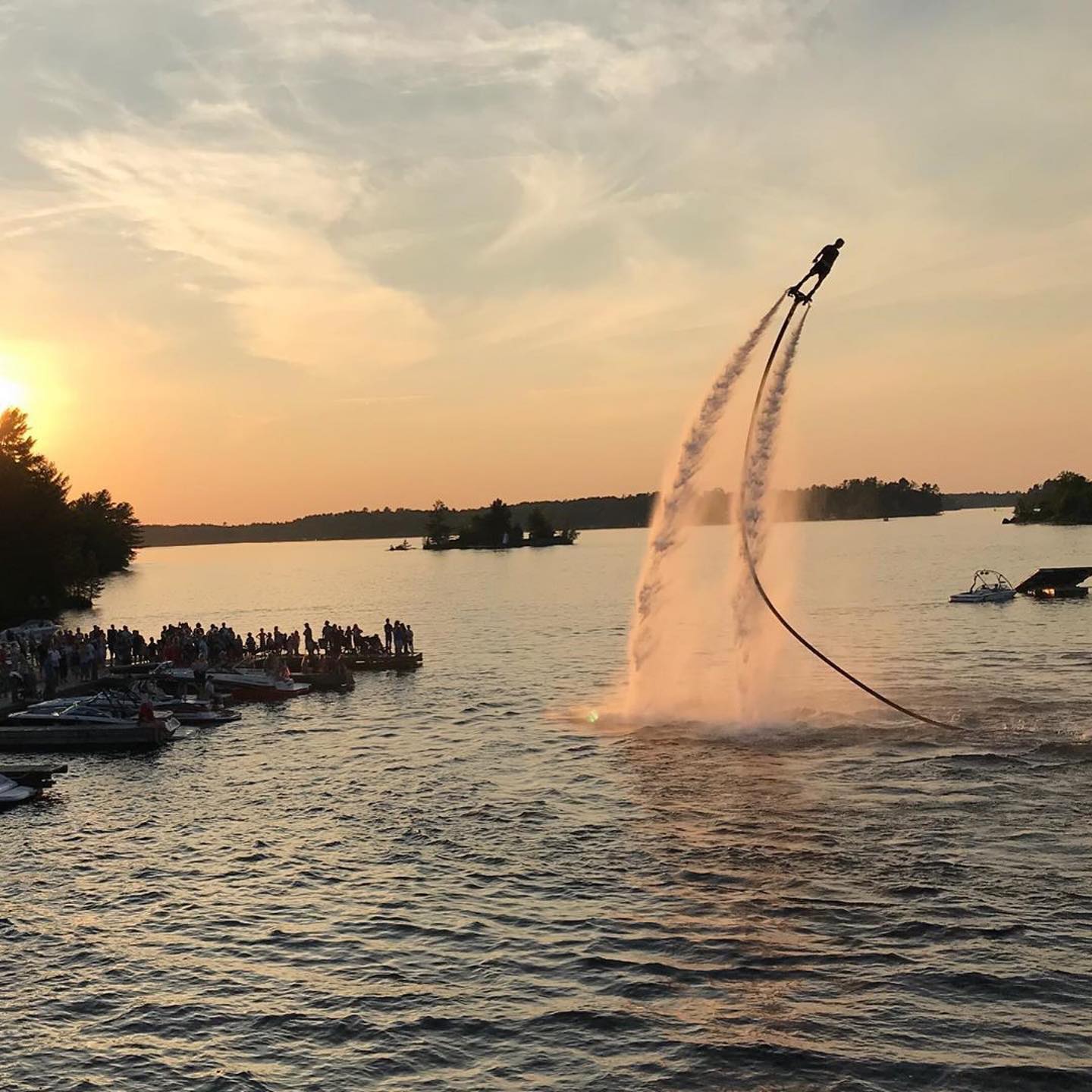 a flyboarder soars high above a lake at sunset, shooting a huge stream of water in their wake. A crowd on the docks below watches and cheers.