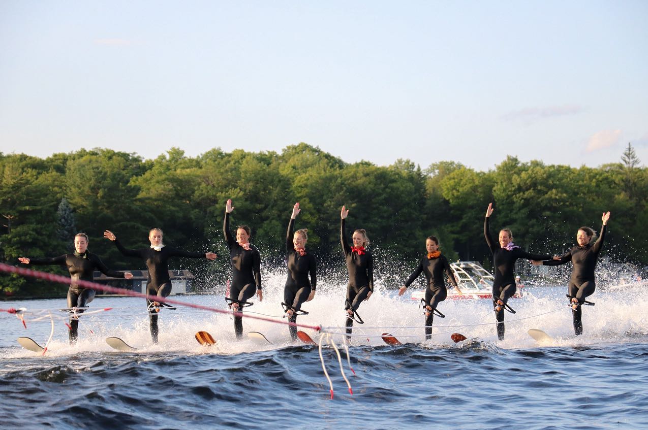 a line of women smile and wave as they waterski on one foot, holding the tow ropes with their other feet.