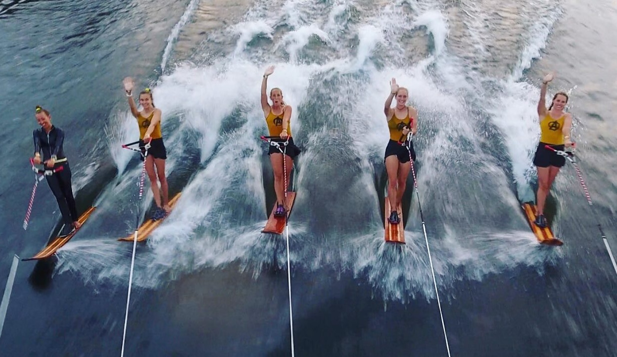 a line of smiling women wave as they wakeboard behind a boat.