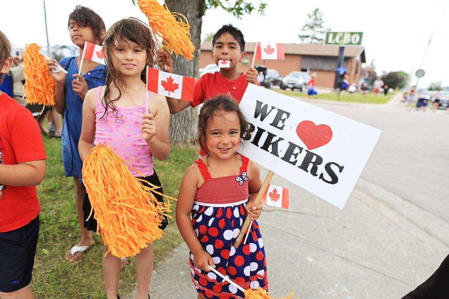 a group of children stand at the side of a road in New Liskeard, waving pom poms and carrying Canada flags and signs that say "We love bikers".