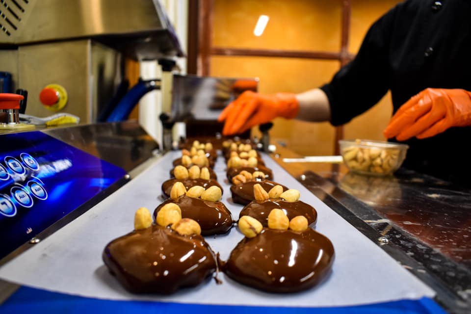a chef wearing gloves garnishes rows of shining, freshly-poured artisan chocolates with nuts.